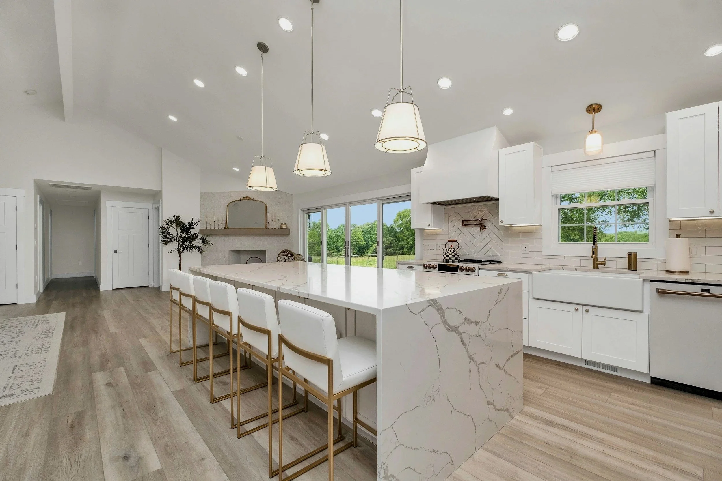 Modern white kitchen with a large marble island, white cabinets, gold fixtures, hardwood floors, with windows showing green trees outside and pendant lights overhead.