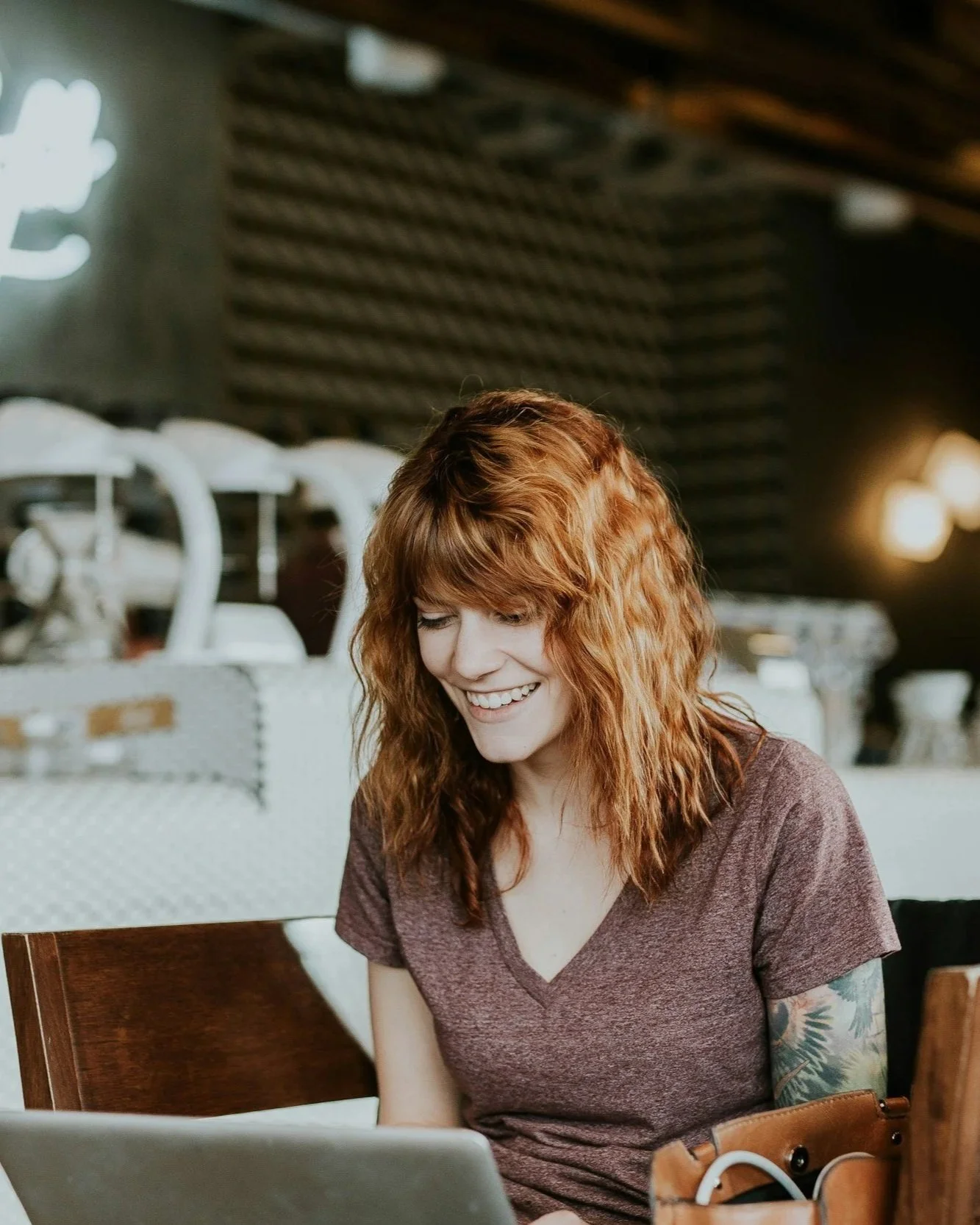 A woman with curly red hair smiling while looking at her laptop in a cafe.