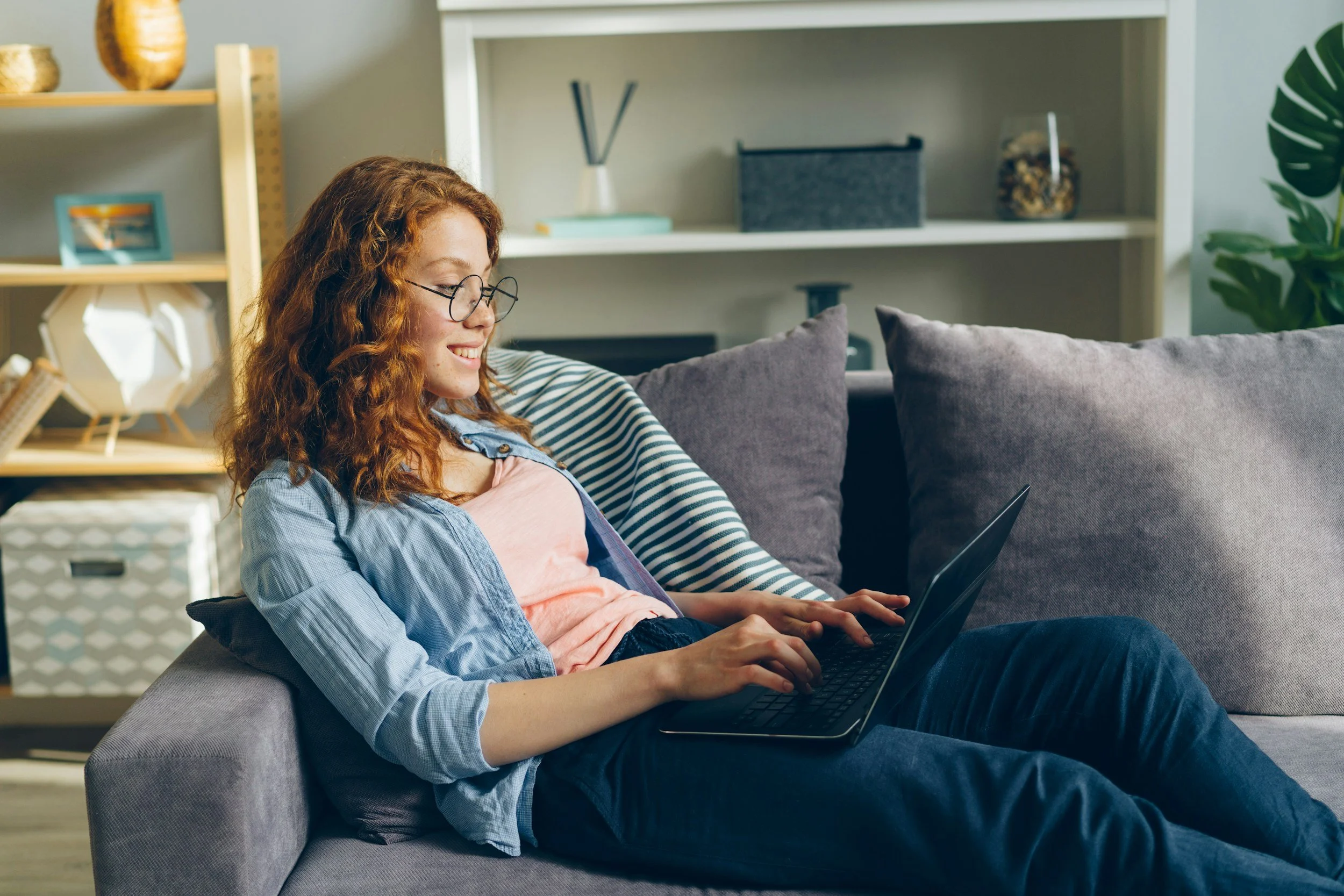 A young woman with curly red hair and glasses is sitting on a gray sofa, working on a laptop. She is smiling and appears relaxed in a cozy living room with wooden shelves, books, decorative items, and green plants.