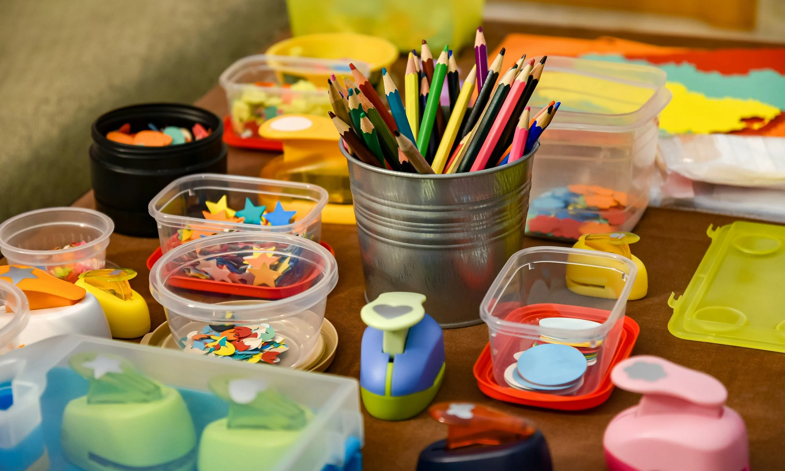 A variety of colorful craft supplies on a table, including colored pencils, star-shaped confetti, paper cut-outs, and small containers filled with craft items, with a metal cup holding colored pencils in the center.
