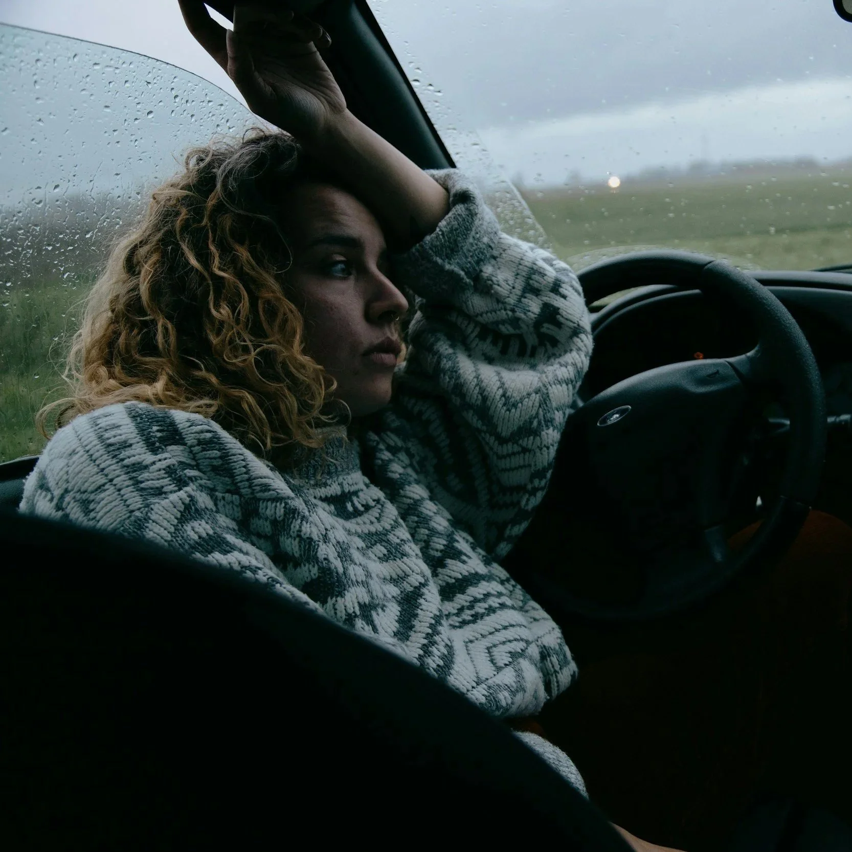 A woman with curly blonde hair sitting in the driver's seat of a car, looking pensively through a rain-covered windshield, with one hand resting on her forehead.