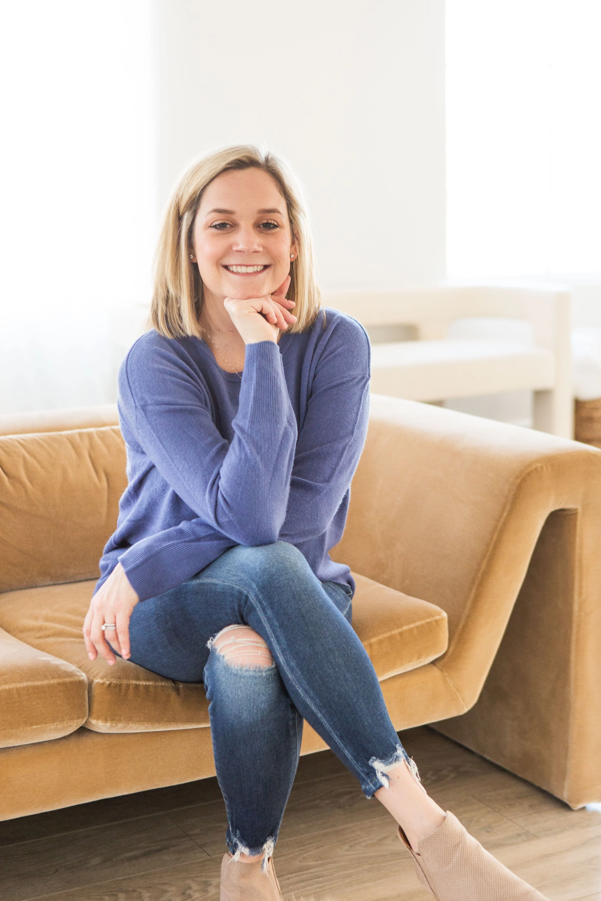 A woman with blonde hair smiling and sitting on a tan couch in a bright room, wearing a blue sweater, ripped jeans, and beige high-heeled shoes.