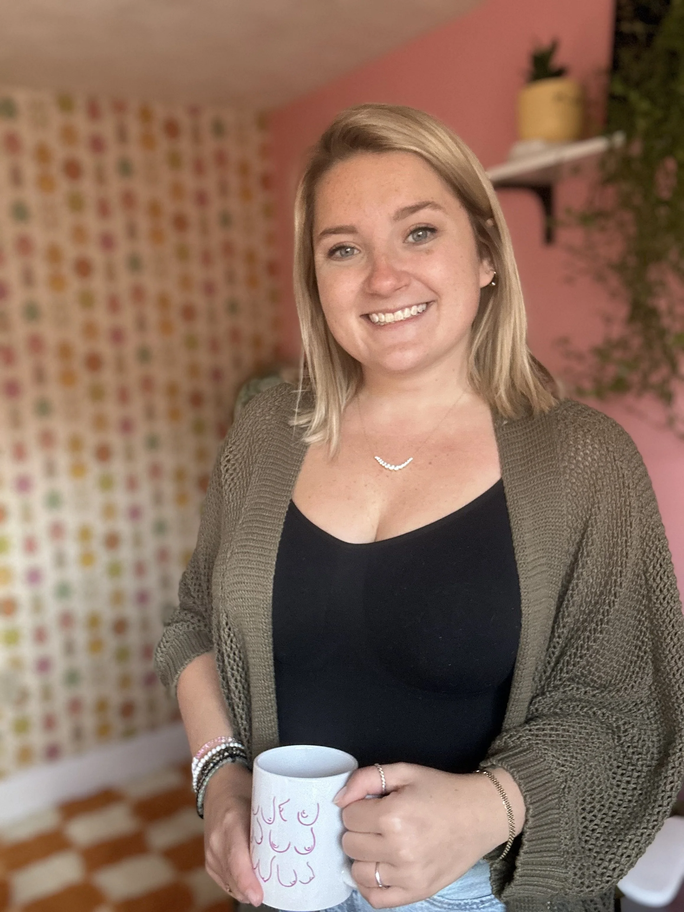 A woman with blonde hair smiling, holding a white mug, standing in a colorful, cozy room with pink walls and patterned wallpaper.