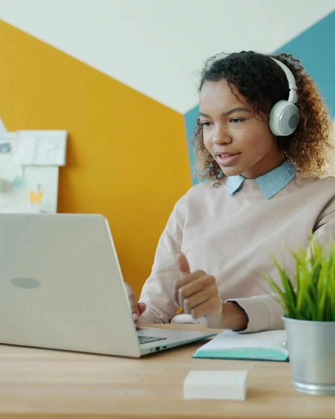 A woman with curly hair wearing headphones, sitting at a desk with a laptop, a notebook, and a potted plant, in a colorful room with yellow, white, and blue walls.