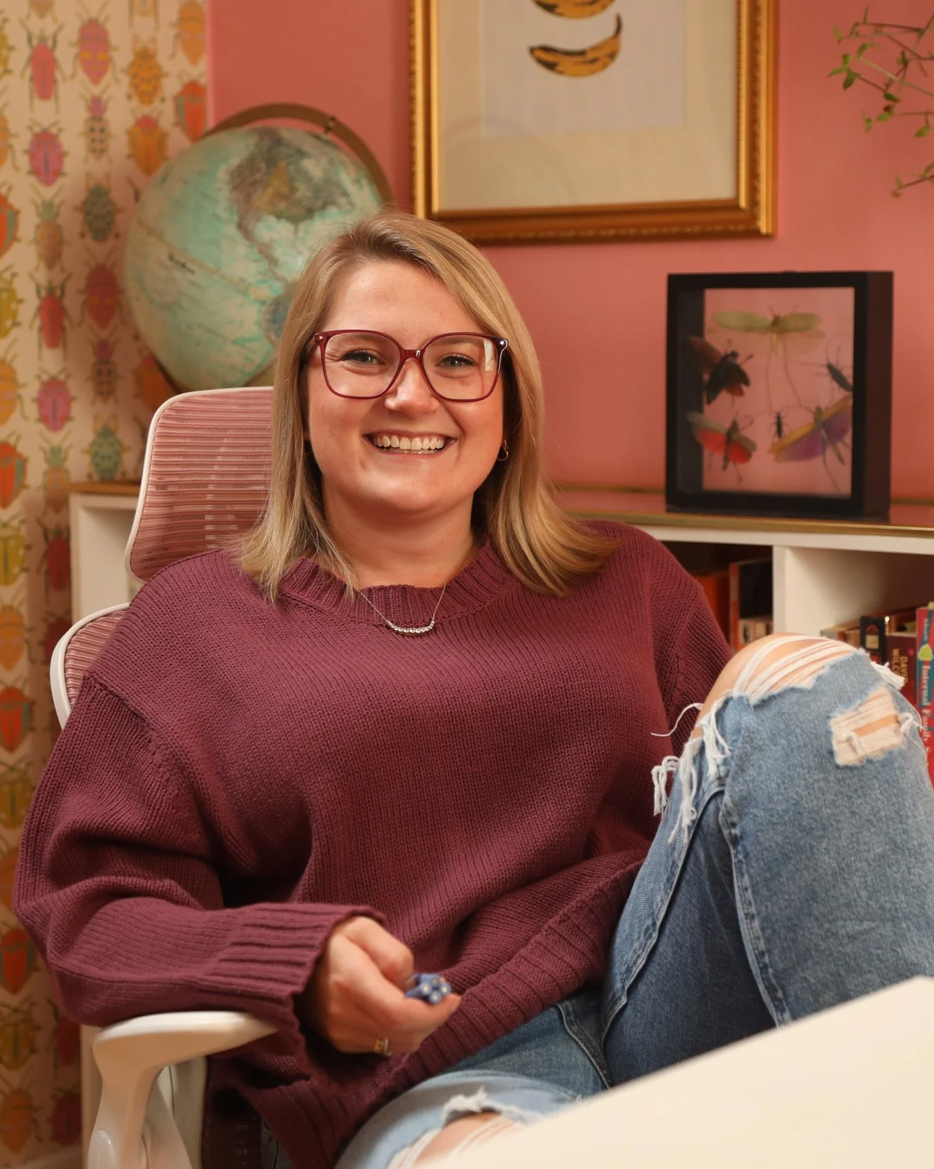A young woman with blonde hair, glasses, and a burgundy sweater sitting in a pink-colored room, smiling at the camera. In the background, there is a globe, framed artwork with insects, a plant, and books on a nearby shelf.