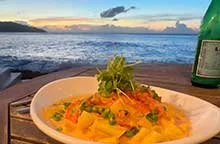 A bowl of seafood pasta garnished with herbs on a wooden table on the water during sunset at Ama.