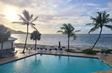 Poolside view at Divi Carina Bay overlooking the ocean with palm trees, lounge chairs, and a cloudy sky.