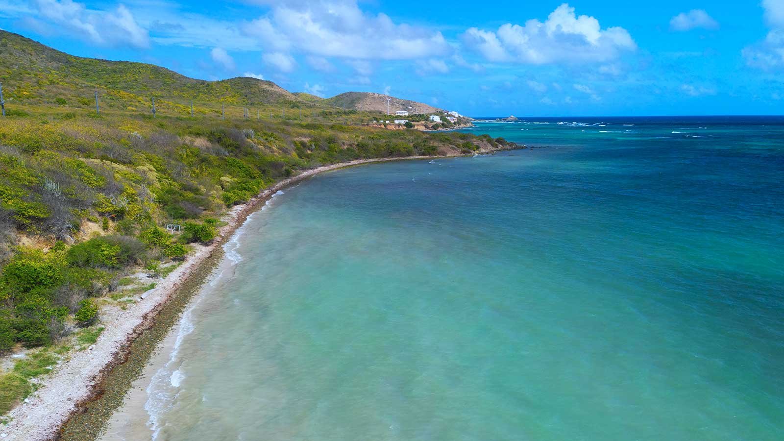 A coastal scene with a sandy beach, clear turquoise water, green hillside, and a partly cloudy blue sky.