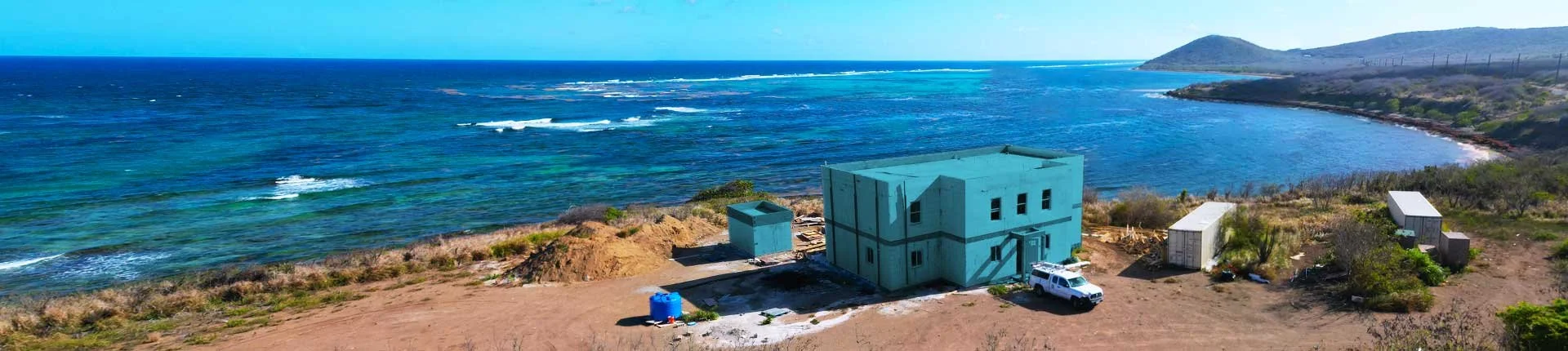 A house under construction on a coastal hillside with the ocean and a distant hill in the background.