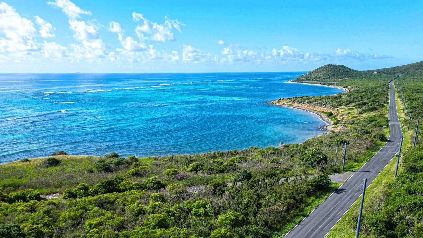 A coastal landscape with a winding road alongside green hills and a blue ocean extending to the horizon, under a partly cloudy sky.