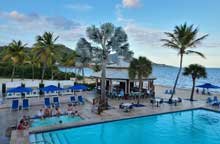 Swimming pool area at Divi Carina Bay resort with lounge chairs, umbrellas, palm trees, and a view of the water in the background.