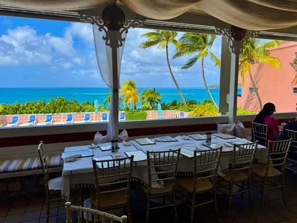 A dining table at Goat Soup and Whiskey set for a meal with white tablecloth, surrounded by wooden chairs, overlooking a tropical beach with palm trees, blue sky, and ocean in the distance.