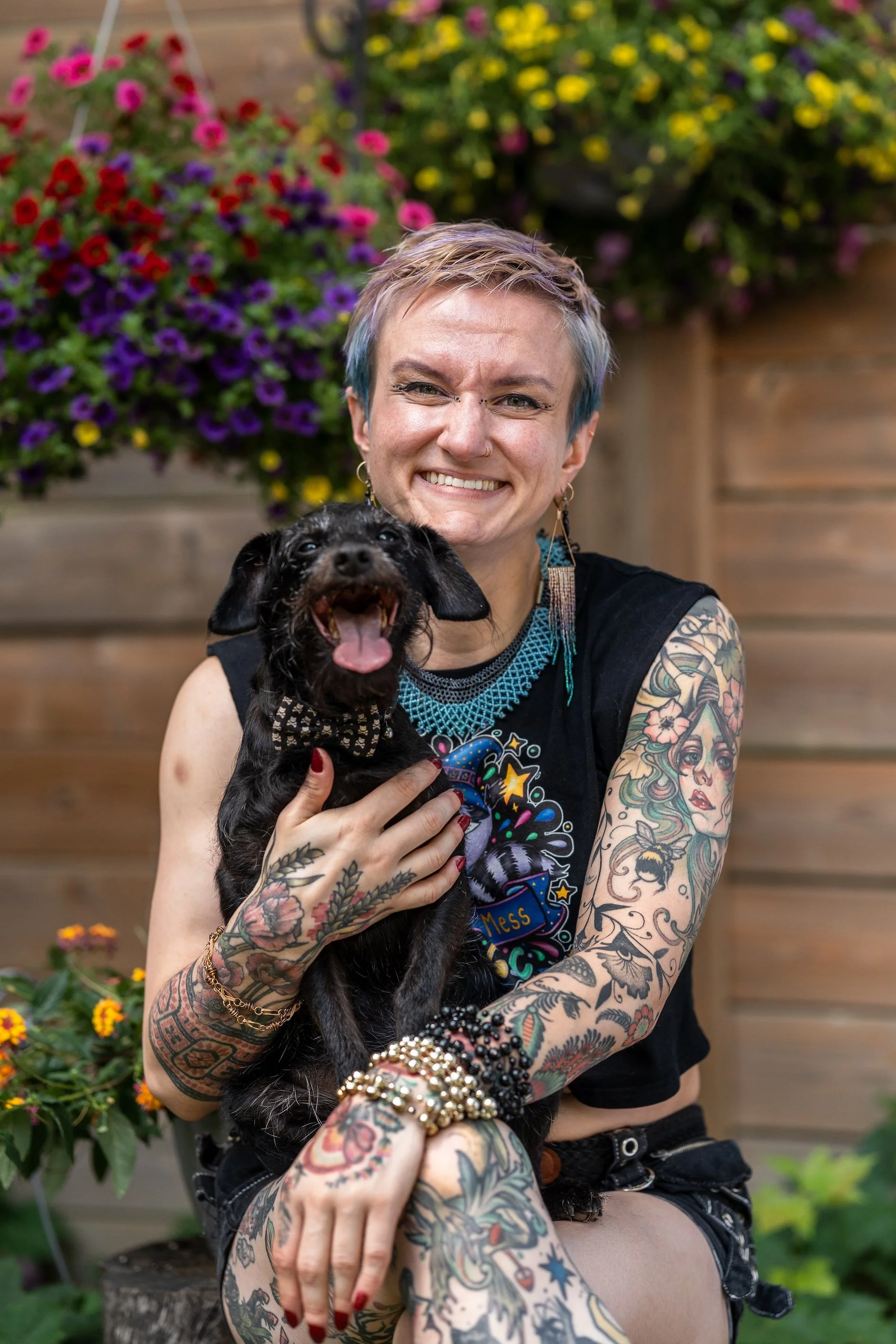 Photo of a smiling person with lots of tattoos holding a smiling little black dog on their lap.