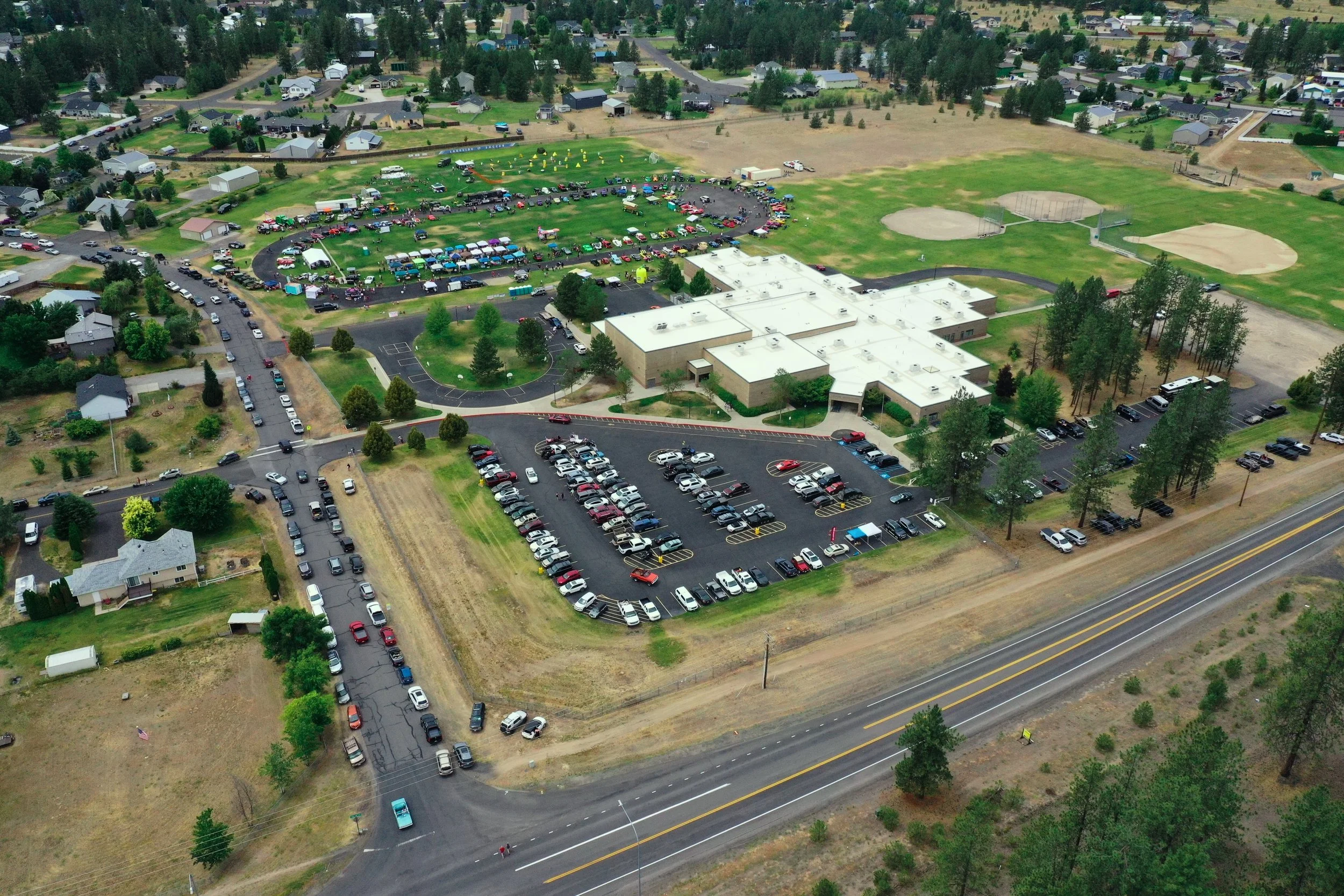 An aerial view of a large community park with many cars parked around the perimeter and inside the park, which features baseball fields, a playground, and a campground with tents. The park is surrounded by residential houses and a highway runs along one side of the park.
