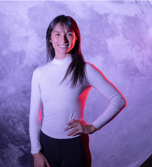 Smile woman in white long-sleeve shirt and black pants standing with hand on hip against textured purple background.
