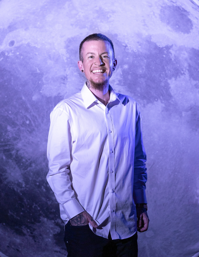 Man smiling in front of a lunar surface backdrop, wearing a white button-up shirt and dark pants.