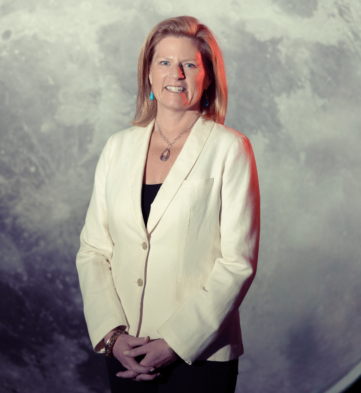 A woman with shoulder-length reddish hair, wearing a white blazer over a black top, smiling slightly with a textured background.