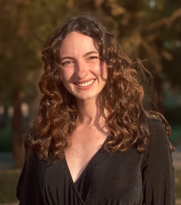 Young woman with curly brown hair smiling outdoors in sunlight, wearing a black top.