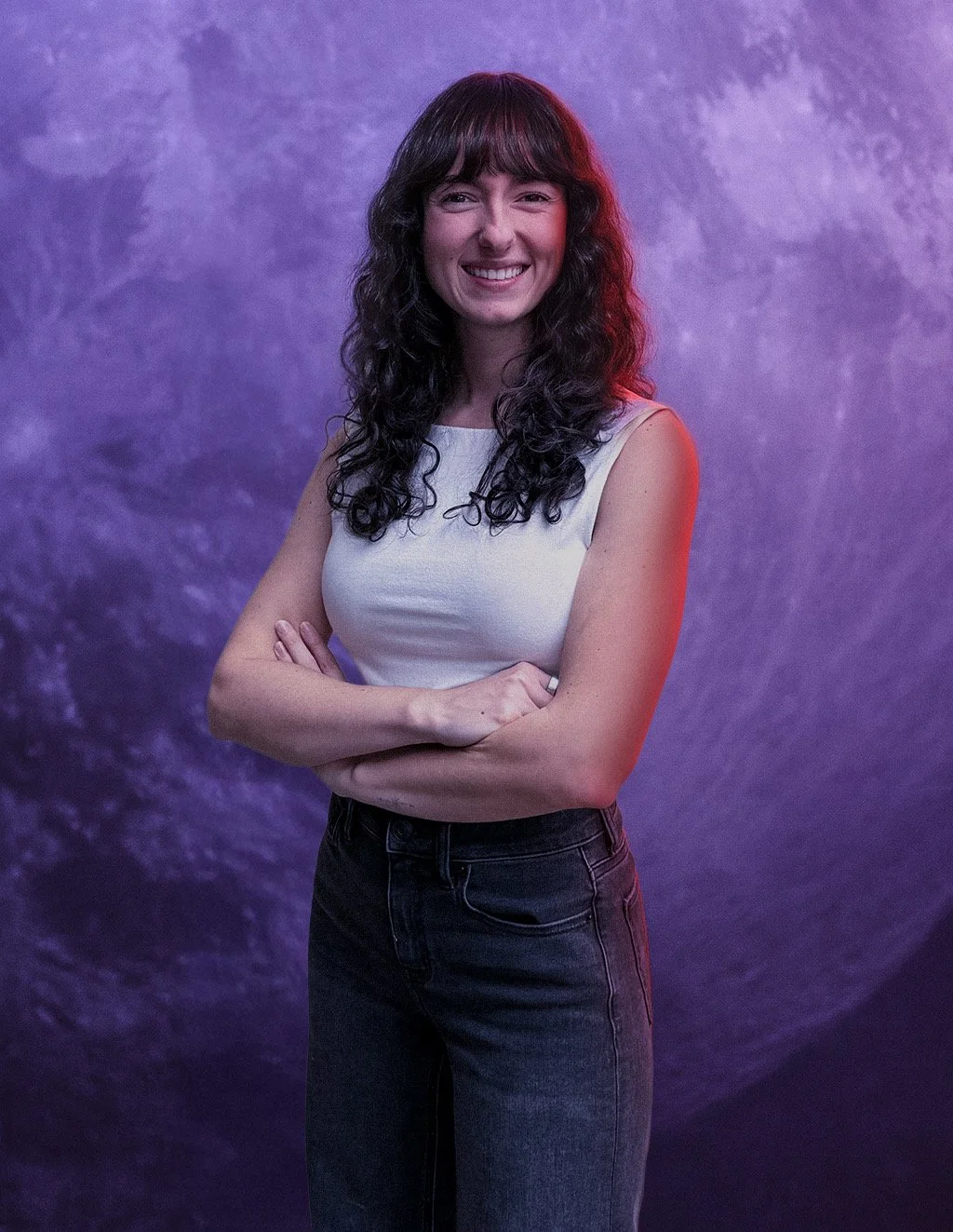 Young woman with curly brown hair smiling outdoors in sunlight, wearing a black top.
