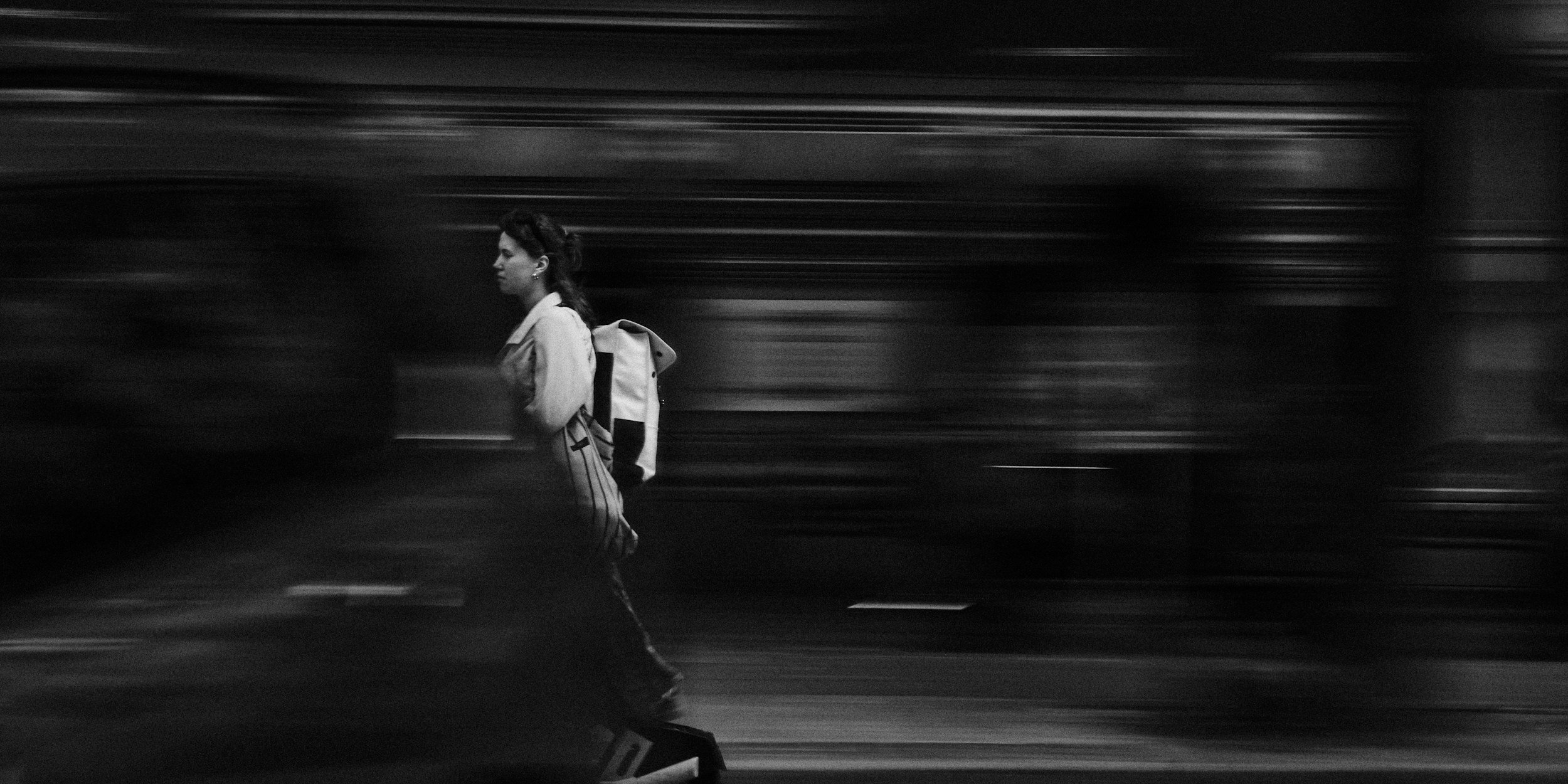 A woman with a backpack walking past a moving train in black and white.
