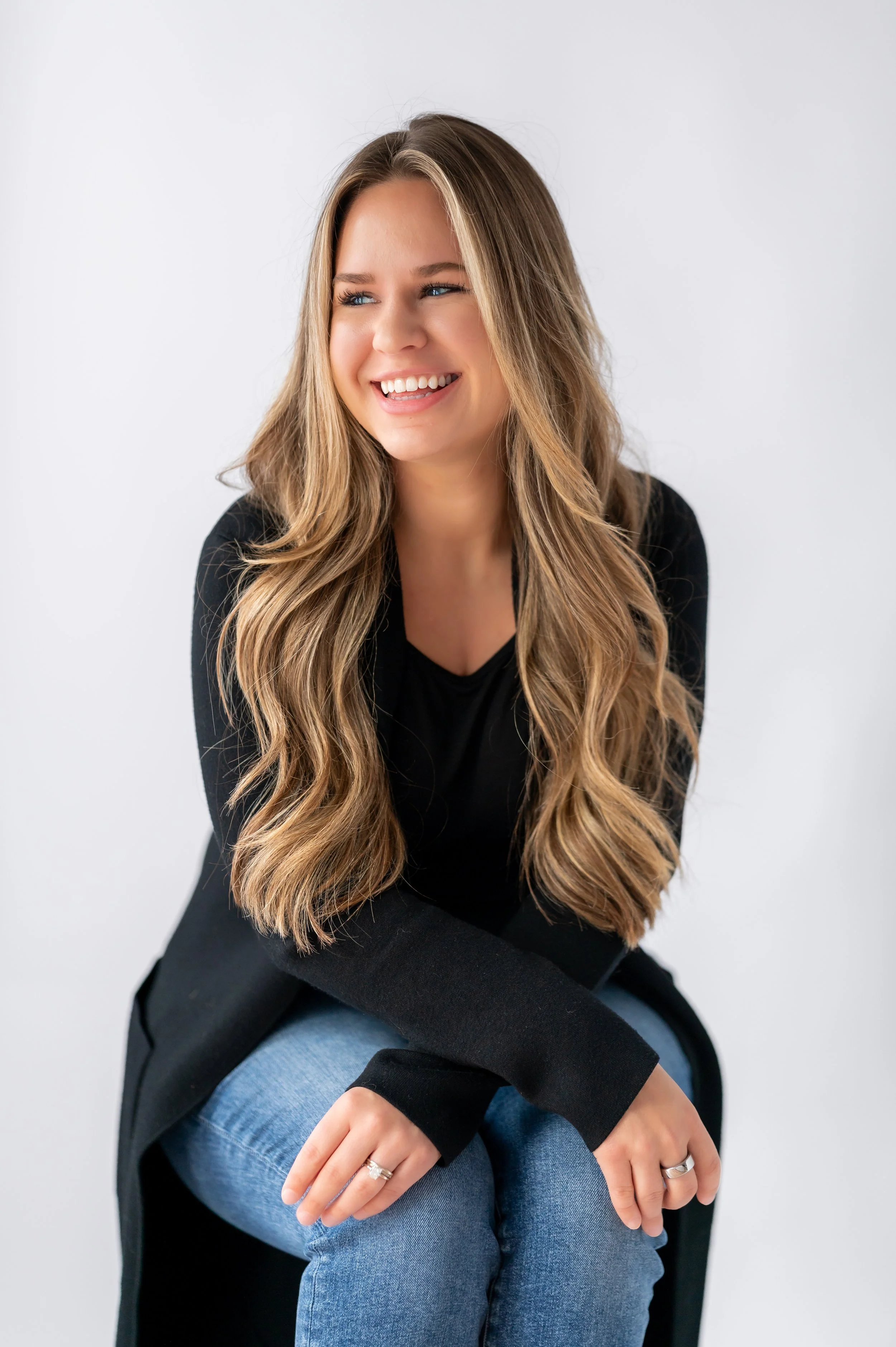 A young woman with long, wavy blonde hair sitting and smiling, wearing a black top and black jacket against a plain white background.