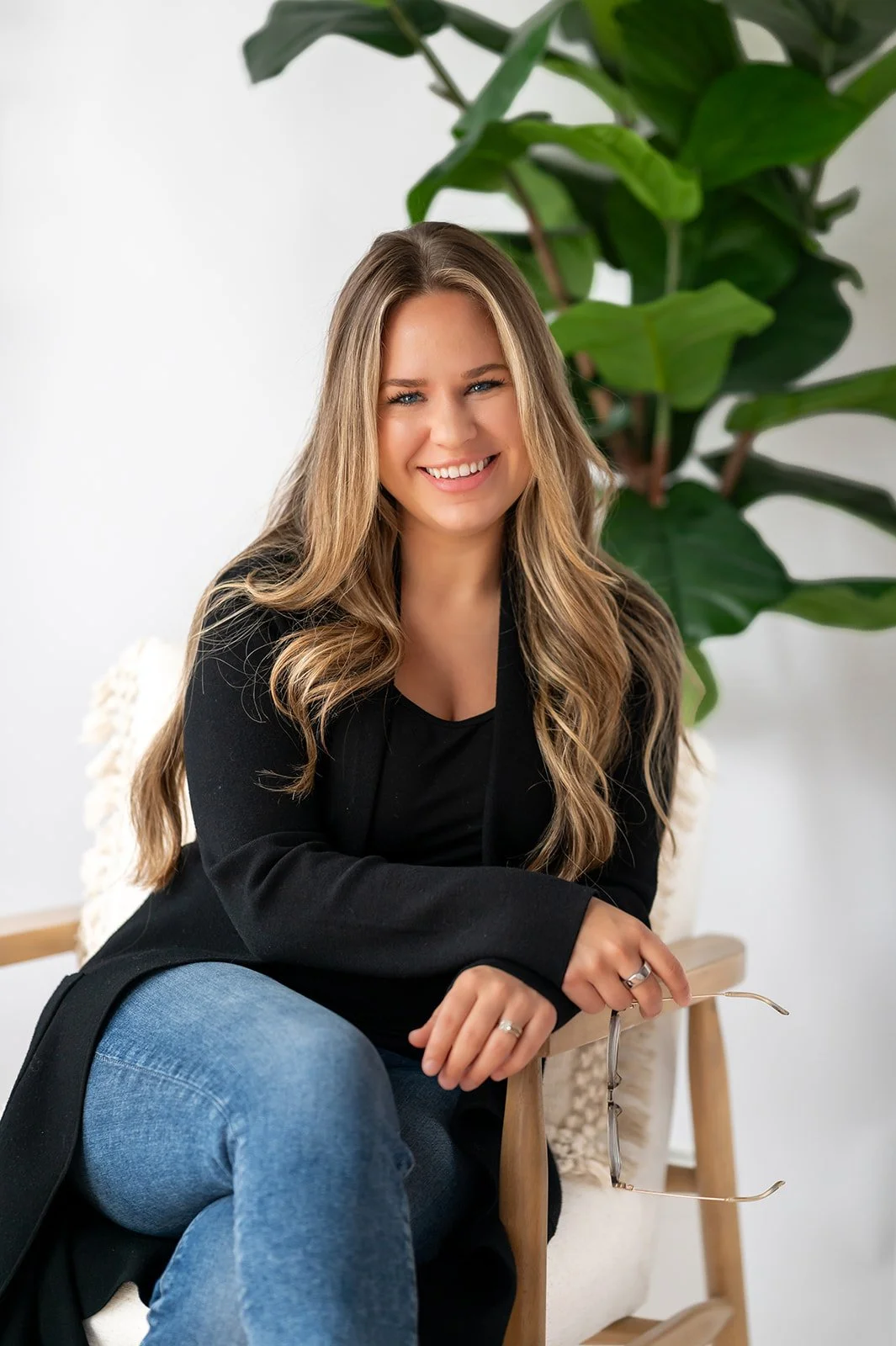 A smiling woman with long, wavy blonde hair sitting on a wooden chair, holding glasses in her right hand, with a large green plant behind her, against a white background.
