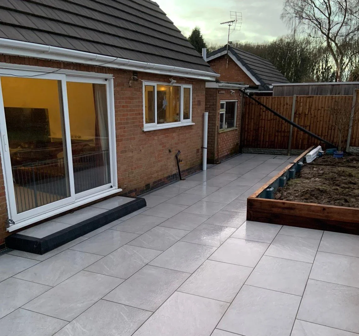 Backyard patio with newly laid light gray tiles, a brick house with sliding glass door and window, wooden fence, and a garden bed with soil, during late afternoon or early evening.
