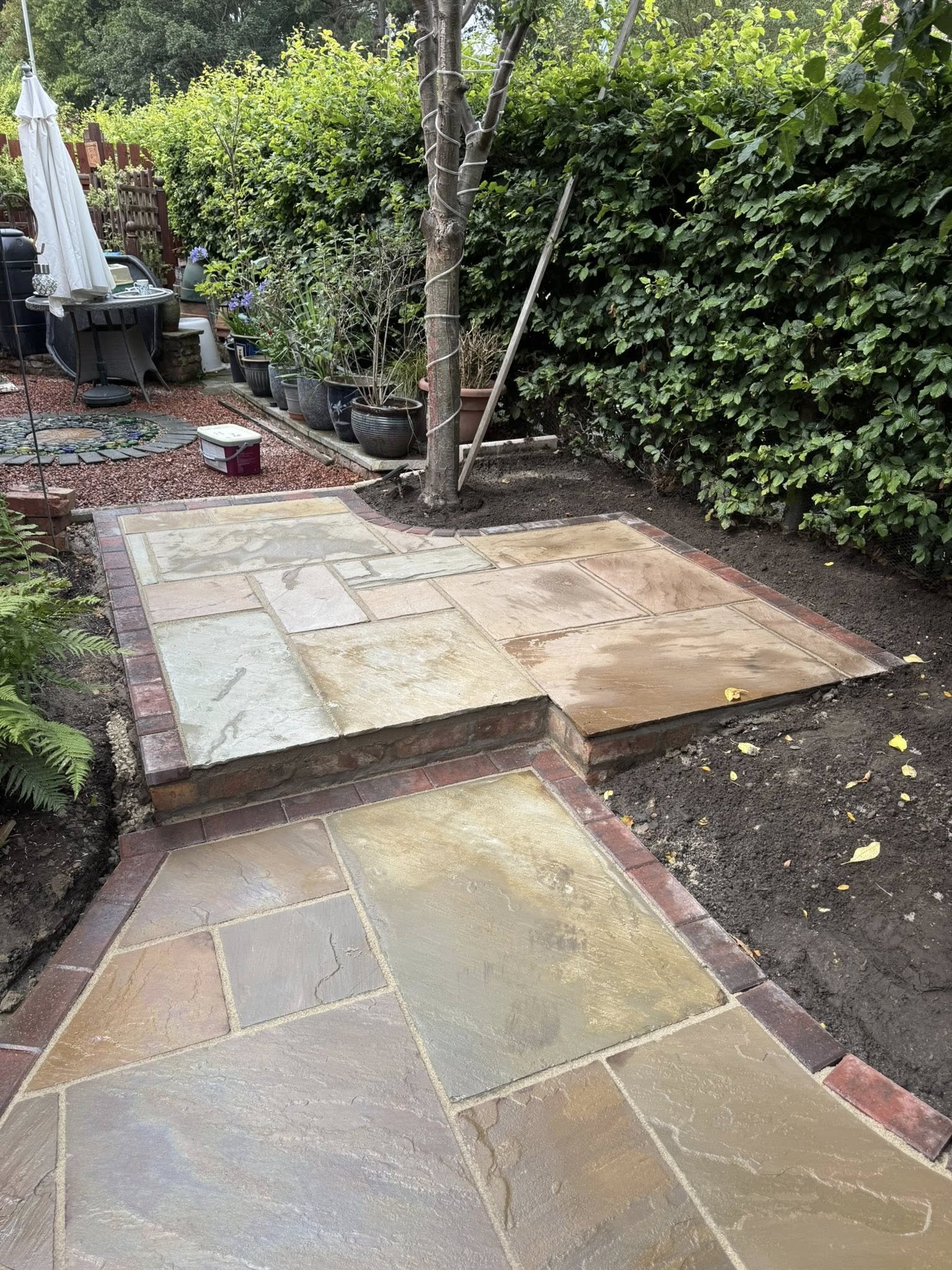 Newly laid stone patio with a small step, surrounded by plants, potted trees, and garden furniture in a backyard.