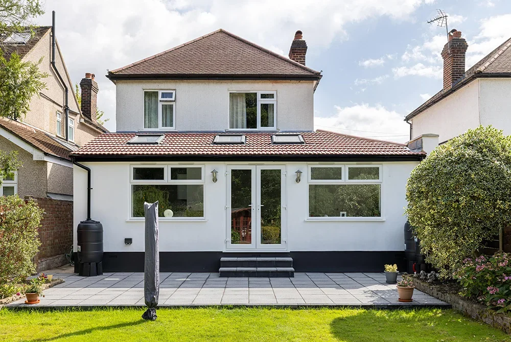 Rear view of a two-story house with white exterior, red tile roof, and a patio with potted plants and outdoor furniture.