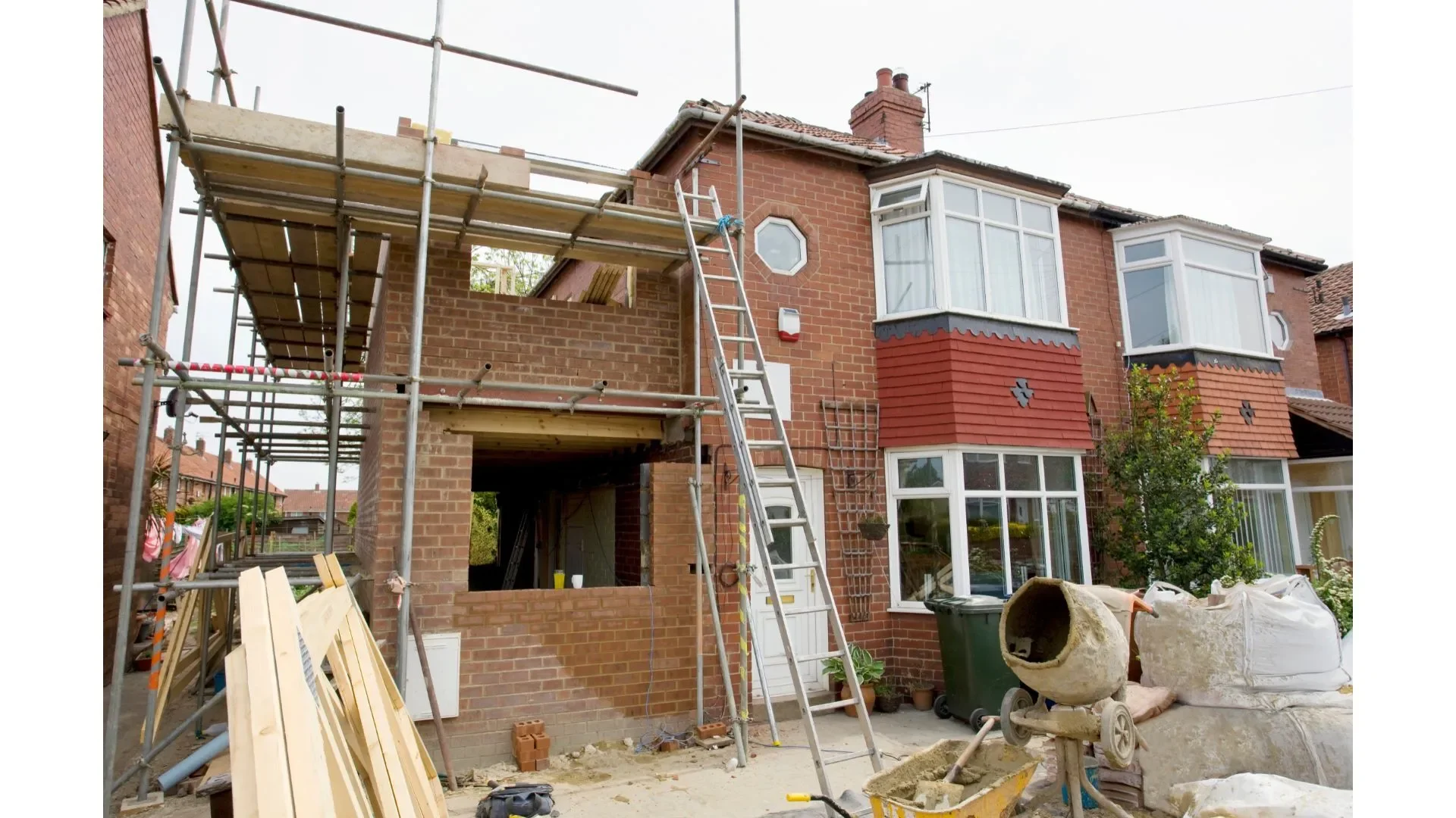 Construction work on a brick house, scaffolding set up in the front, with construction materials and tools around, including a cement mixer, ladder, and wooden planks.