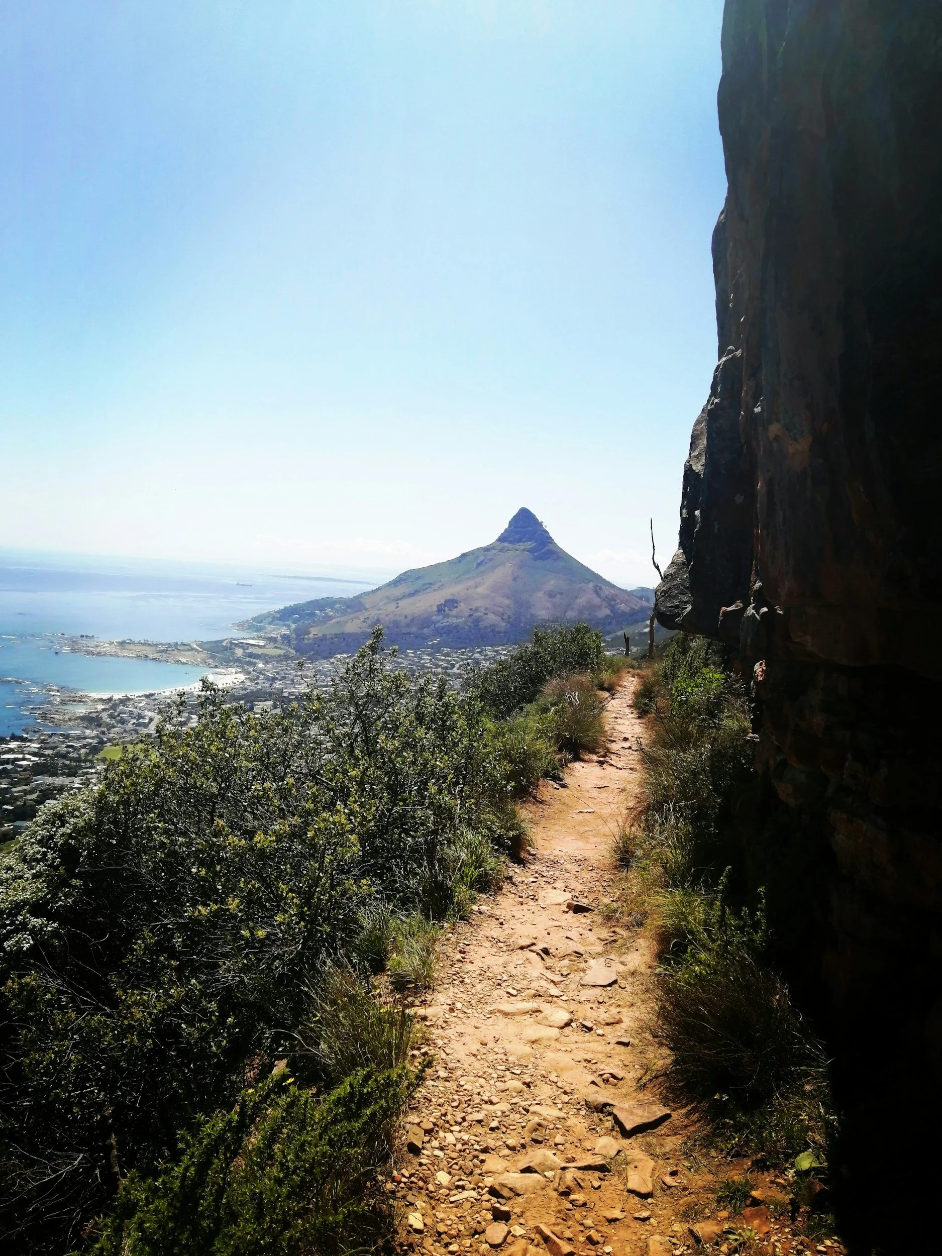 A narrow dirt trail winding along a hillside with greenery, overlooking a bay and a mountain with a pointed peak under a bright blue sky.