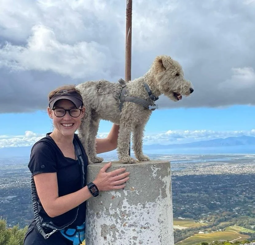 A woman smiling in outdoor attire with a dog standing on a concrete pillar at a high elevation during daytime with cloudy skies.