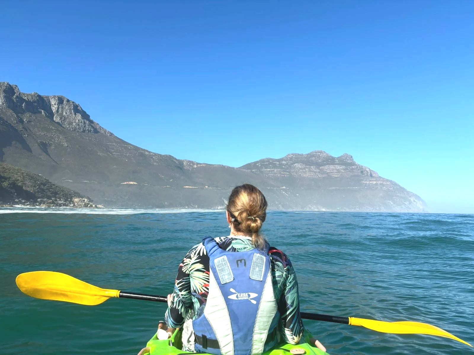 A woman kayaking on a body of water, with mountain cliffs in the background under a clear blue sky.