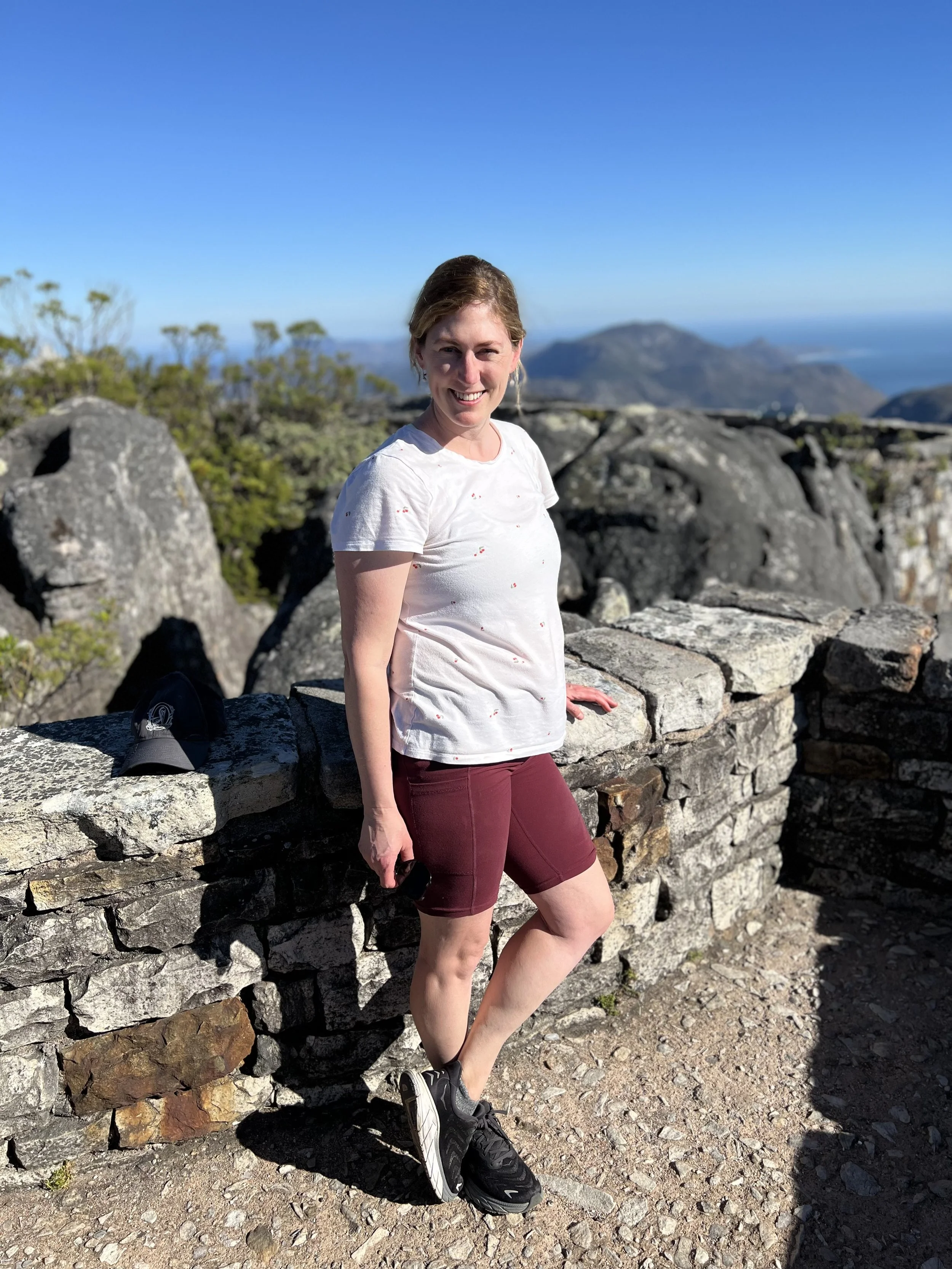 A woman standing outdoors on a stone wall in a mountainous area with clear blue sky, wearing a white T-shirt, maroon shorts, and running shoes, smiling at the camera.