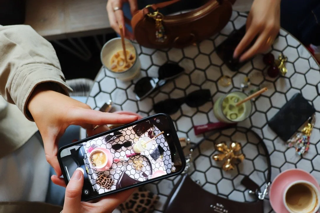 A person taking a photo of a table with various drinks, sunglasses, and jewelry using a smartphone.