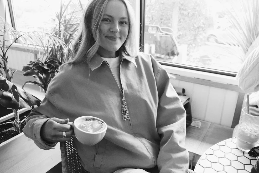 A woman with long hair smiling while holding a coffee cup inside a cafe with plants and a window behind her.