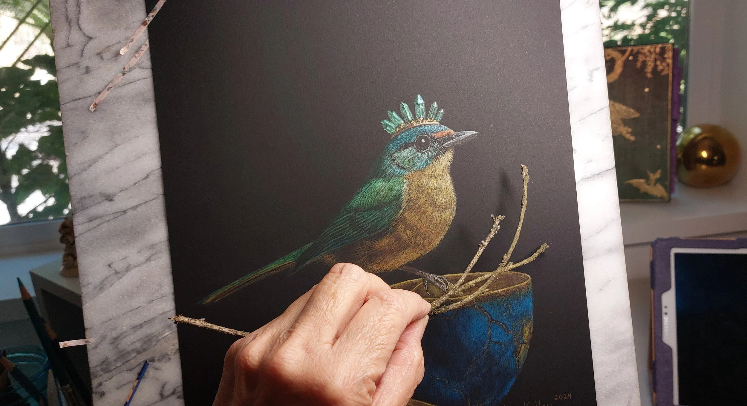 The unfinished drawing for Stir The Awakened showing an emerald‑crowned green bird perched on a cracked teacup, with artist Kim Kelley’s hand holding real twigs in front of the artwork for reference.