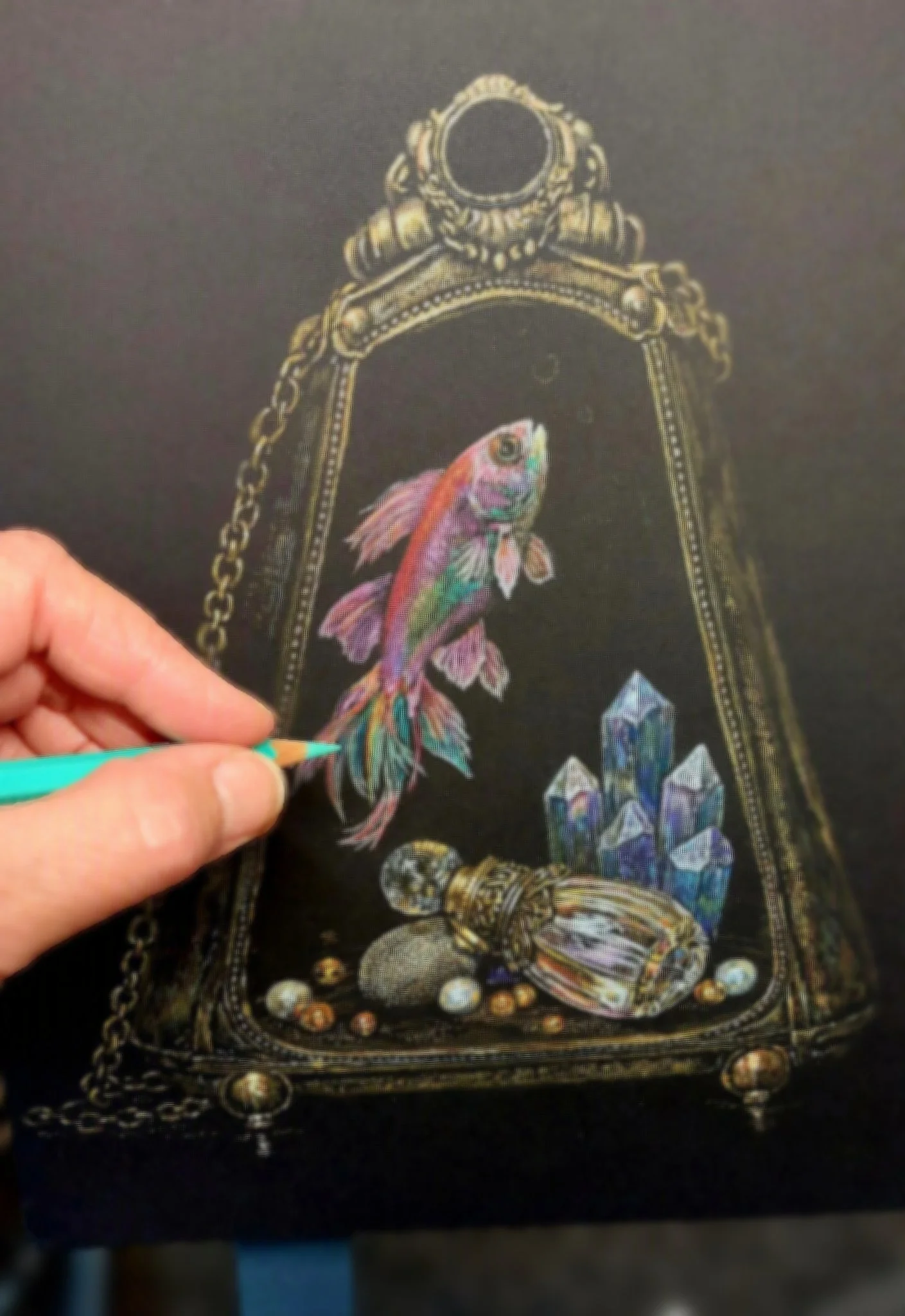 Hand of Artist Kim Kelley working on a colored pencil illustration featuring a fish, purse, perfume bottle, and gemstones