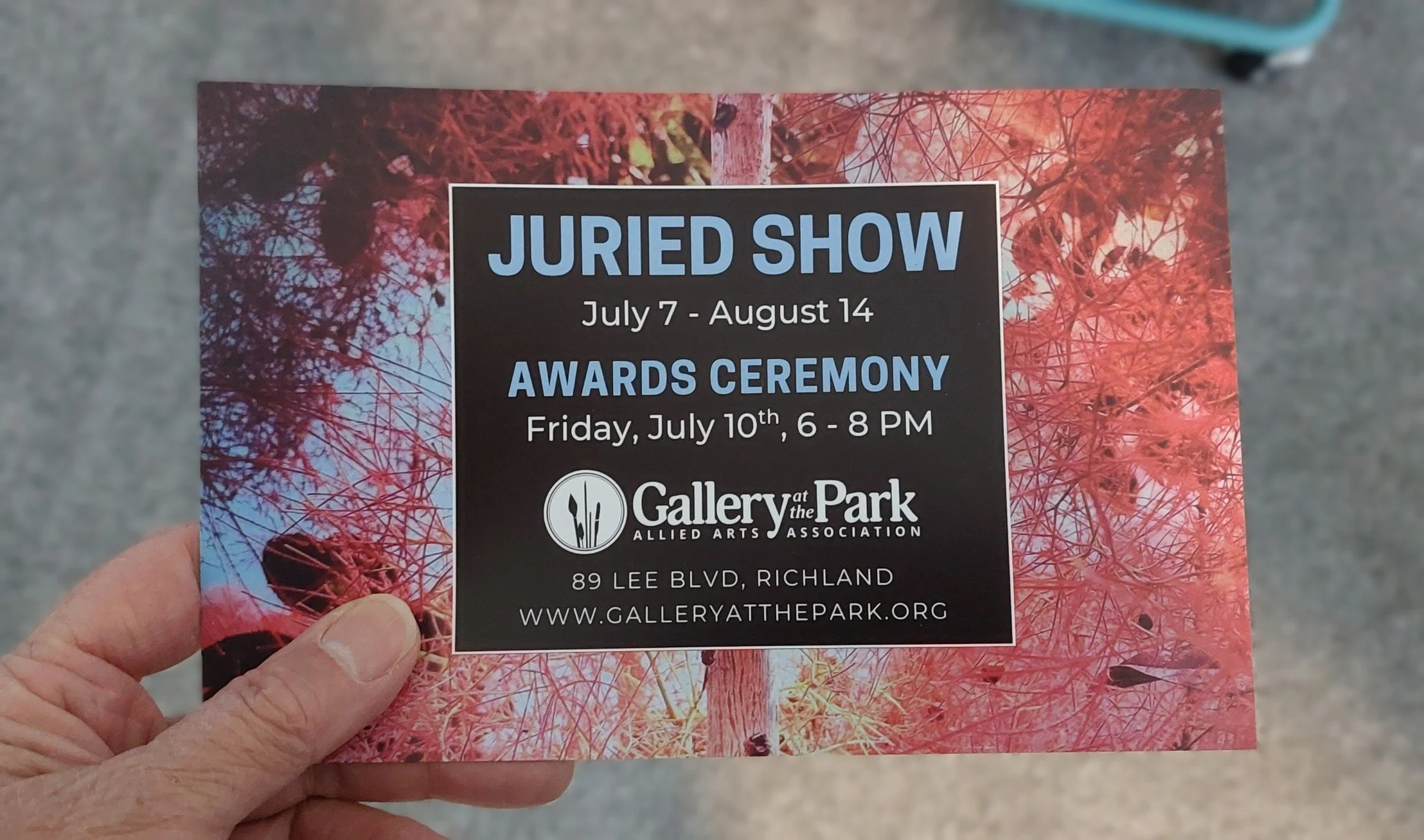 Artist Kim Kelley's hand holding a printed invitation card for the Gallery at the Park Juried Show, with event dates and location visible.