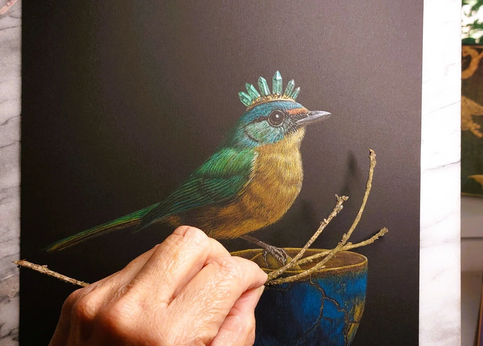Realistic colored pencil drawing of a colorful bird with an emerald crystal crown crest perched on a kintsugi cup with golden egg and branches, held in artist Kim Kelley's  hand.