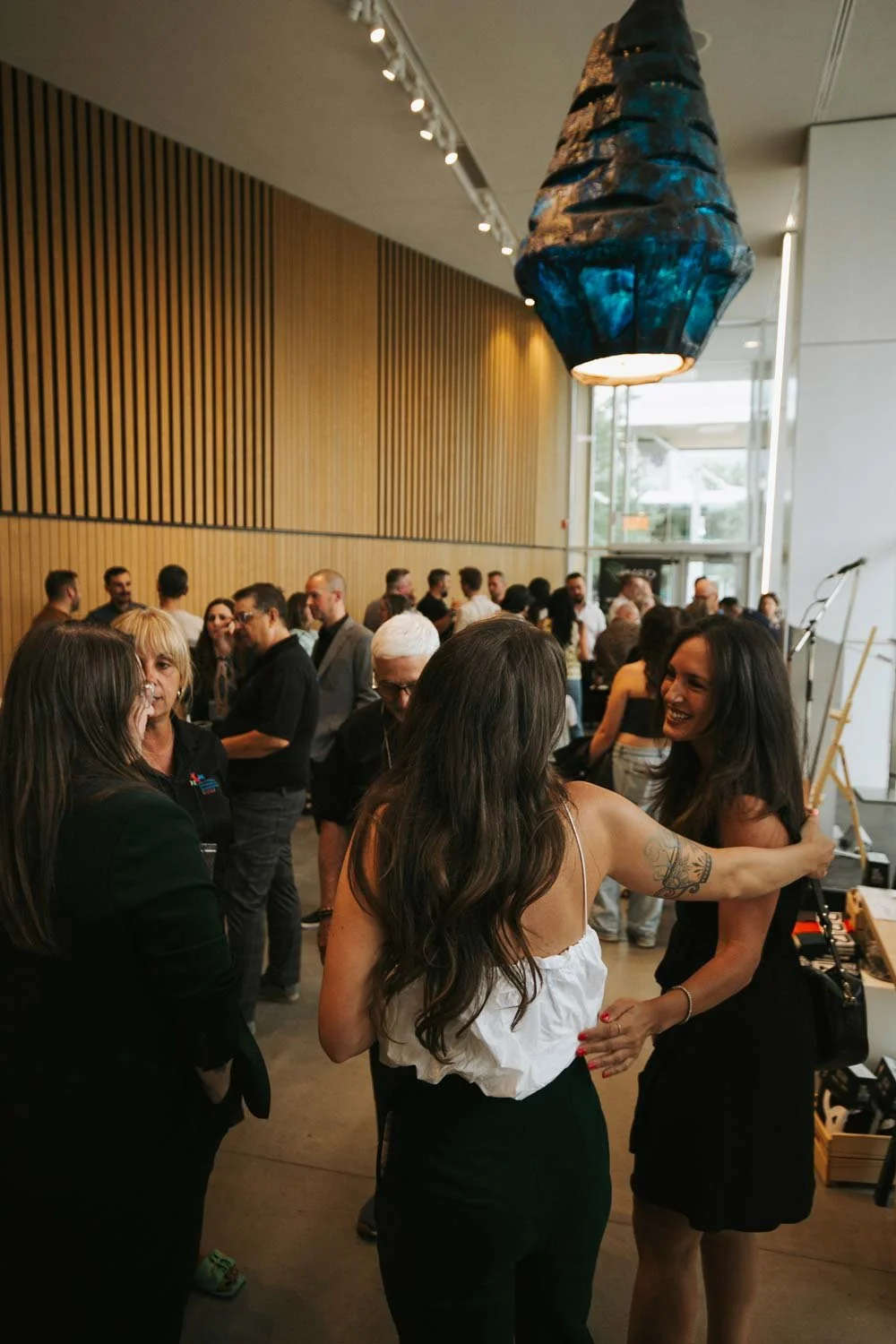 People socializing at an indoor event with a modern wooden panel wall, large window, and decorative hanging light fixture.