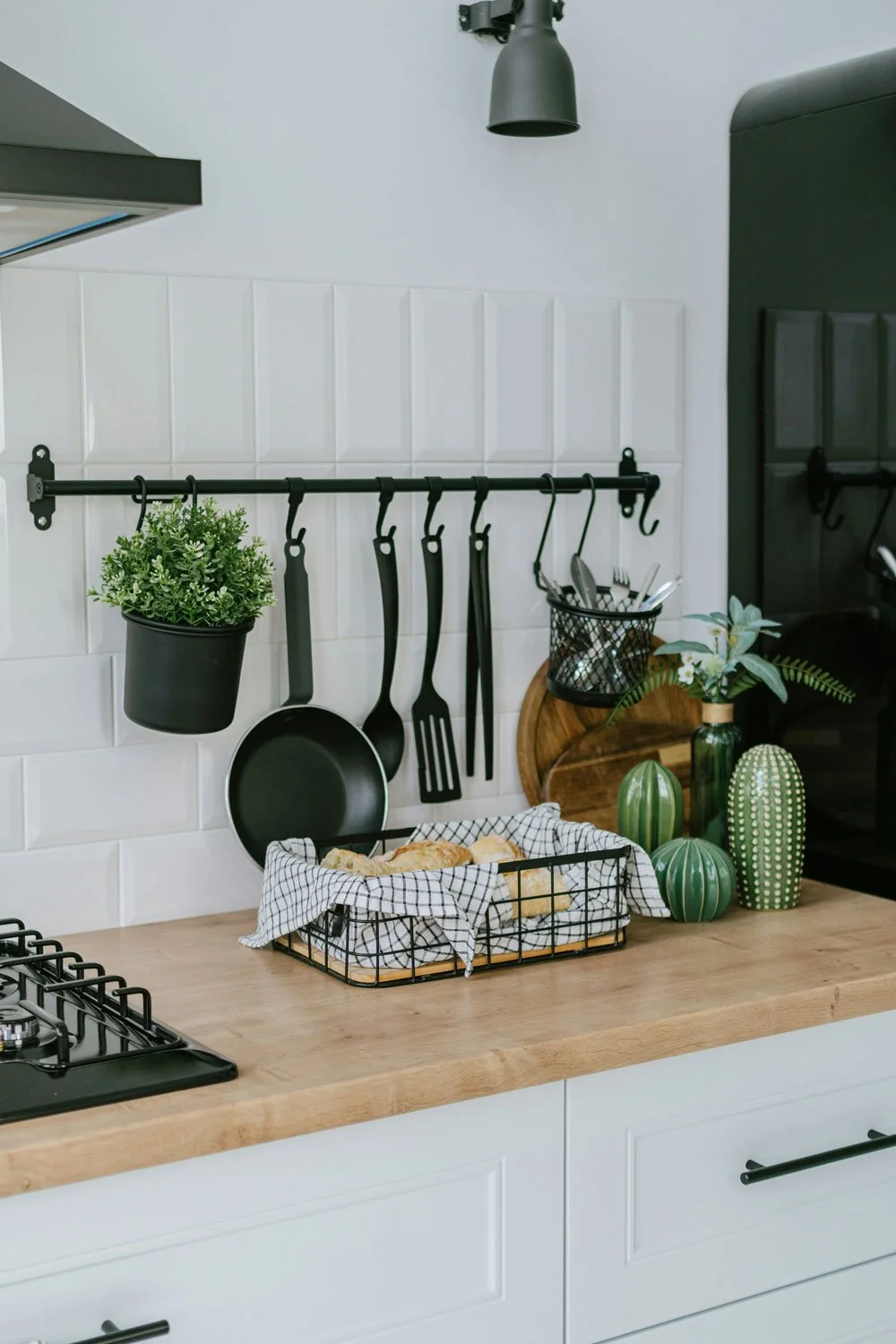 Kitchen counter with a black wire basket holding bread, green and white decorative vases, a black pot with a green plant, a black hanging utensil rack with pots and tools, and a black wall-mounted light fixture.