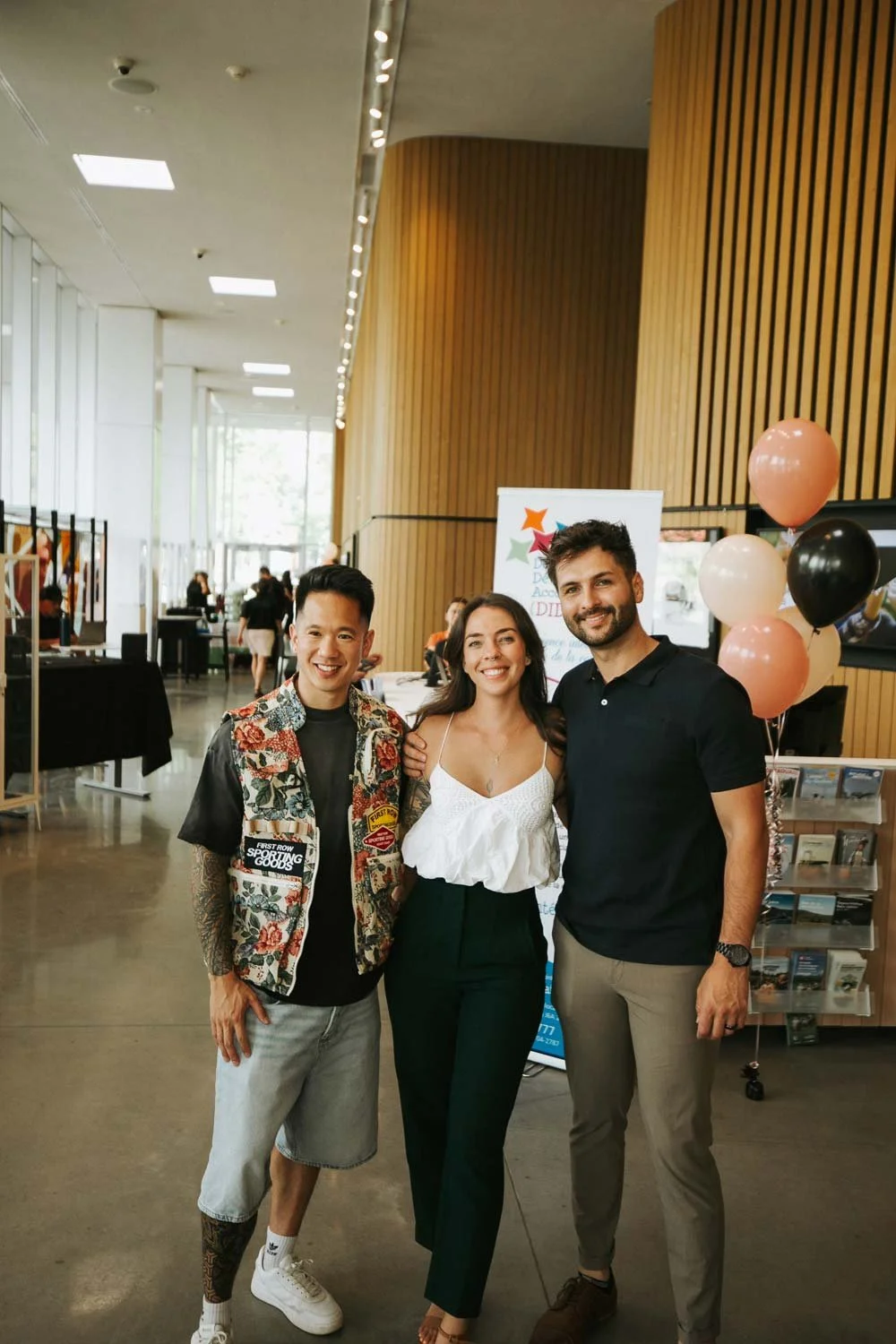 Three people standing indoors at an event, smiling, with balloons and informational display behind them.