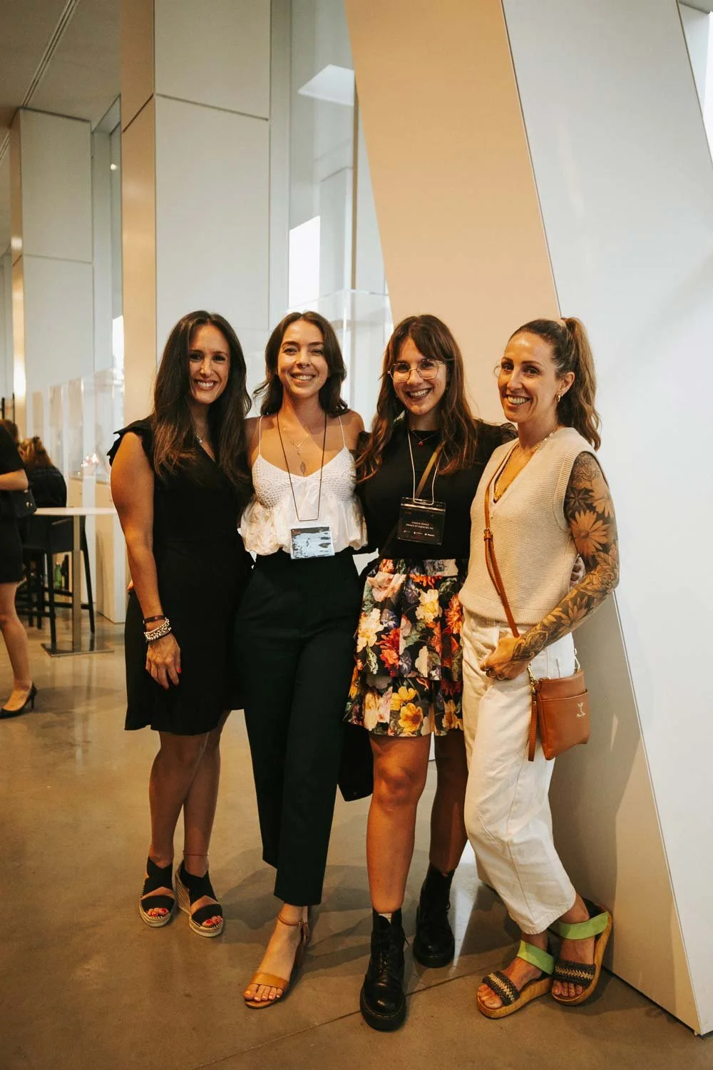 Four women standing together smiling in a modern indoor setting, dressed in stylish casual and semi-formal attire.