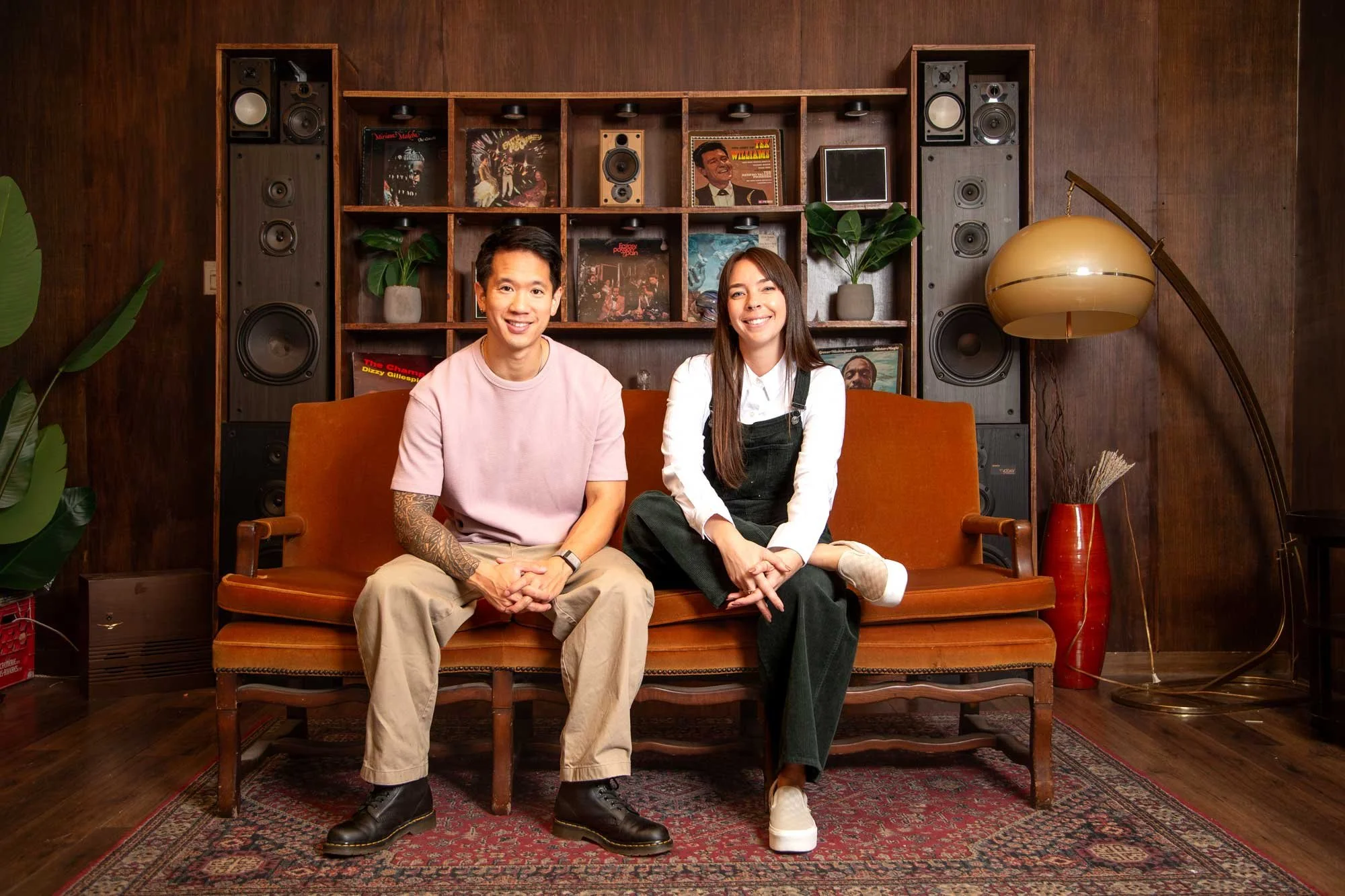 Viet and Ariane sitting on a vintage orange sofa in front of a wooden wall with a bookcase.