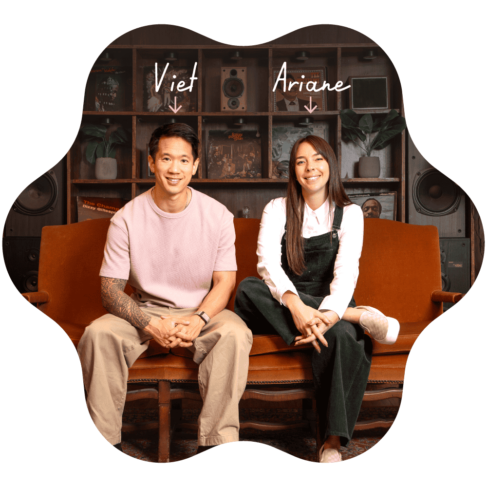 A man and a woman sitting on a brown vintage sofa in a room with wooden shelves, plants, and speakers in the background. The man, labeled "Viet," has short dark hair, a pink T-shirt, beige pants, and a tattoo on his left arm. He is smiling with hands clasped. The woman, labeled "Ariane," has long brown hair, a white shirt under black overalls, and white shoes. She is also smiling, sitting with her legs crossed.