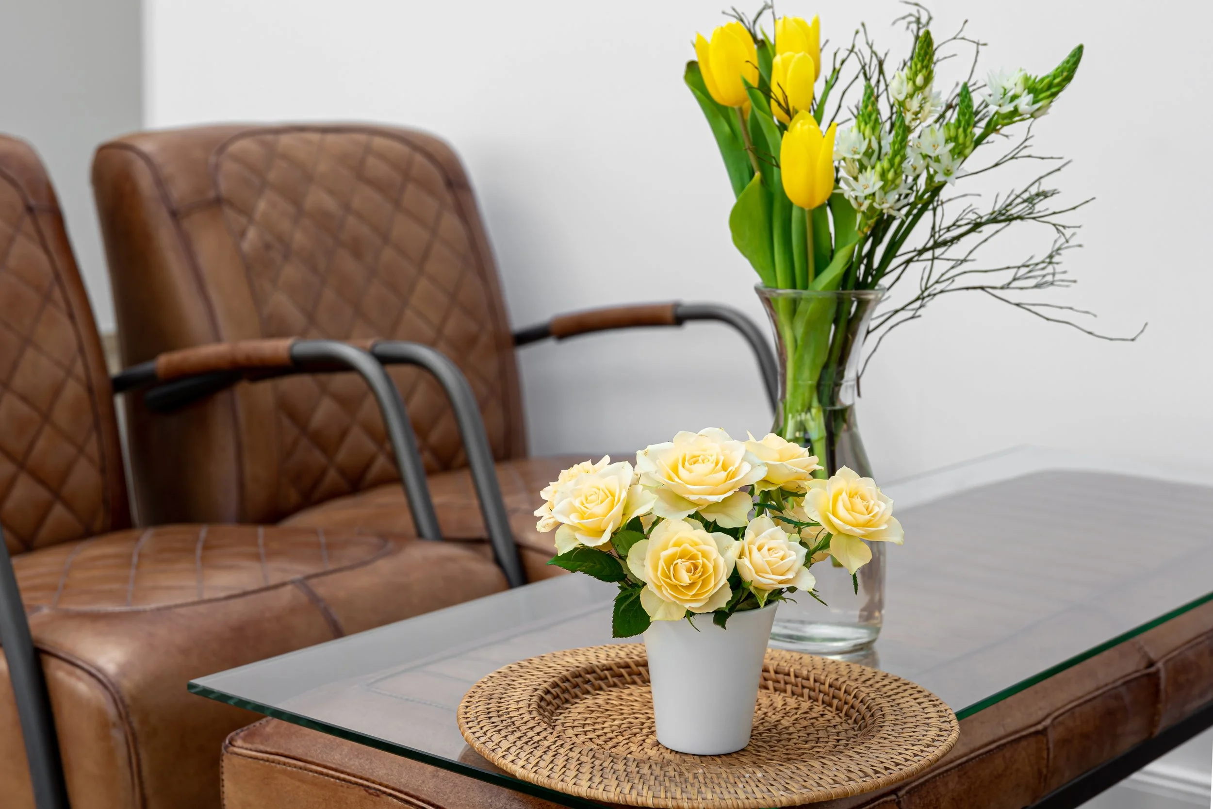 A glass vase with yellow tulips and white flowers, a white flowerpot with cream-colored roses, and a woven placemat on a glass table, with brown leather chairs in the background.