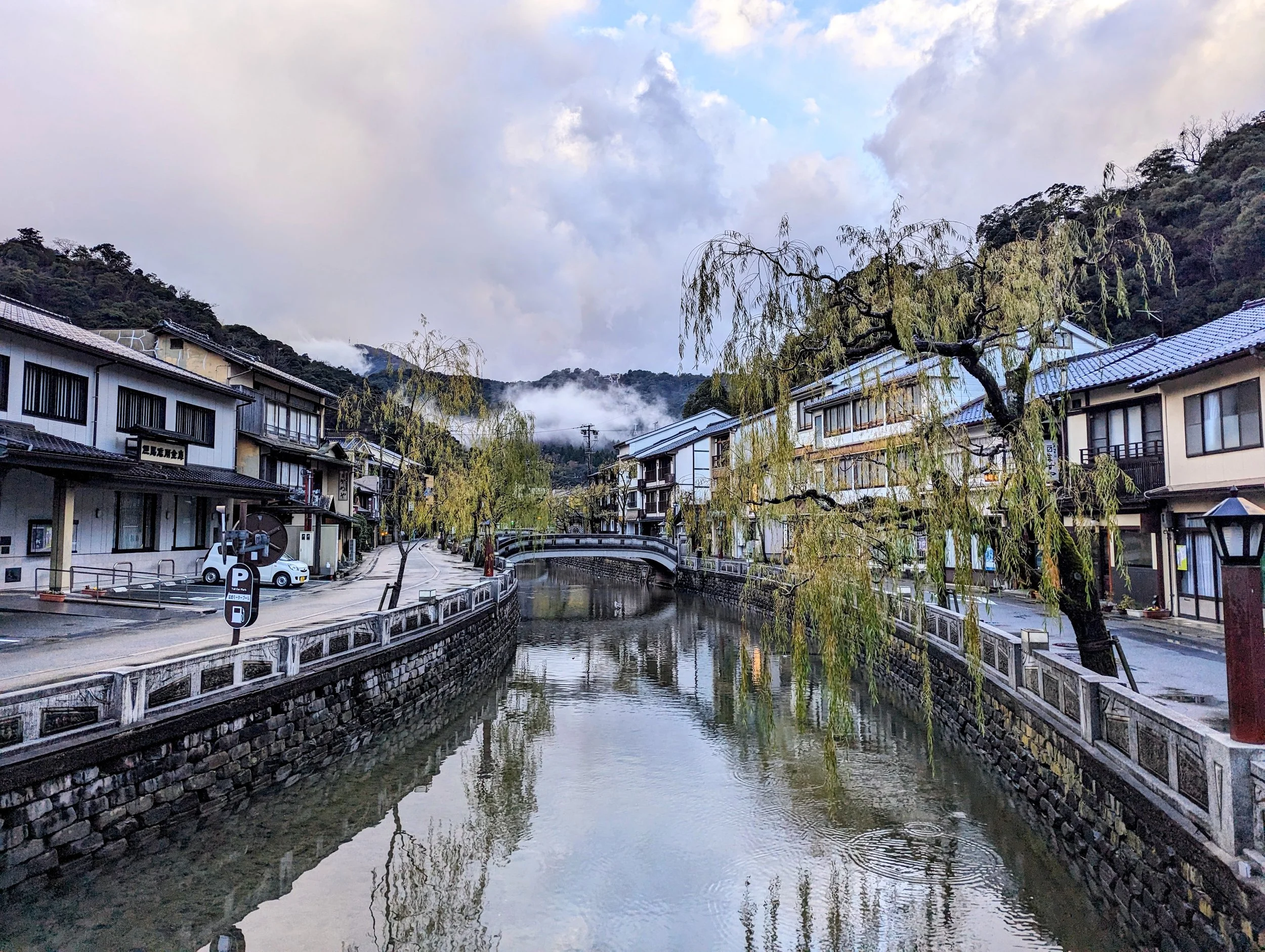A quiet street running along a narrow river, with traditional Japanese buildings on both sides, a bridge over the river, and mist-covered hills in the background, with trees and clouds in the sky.