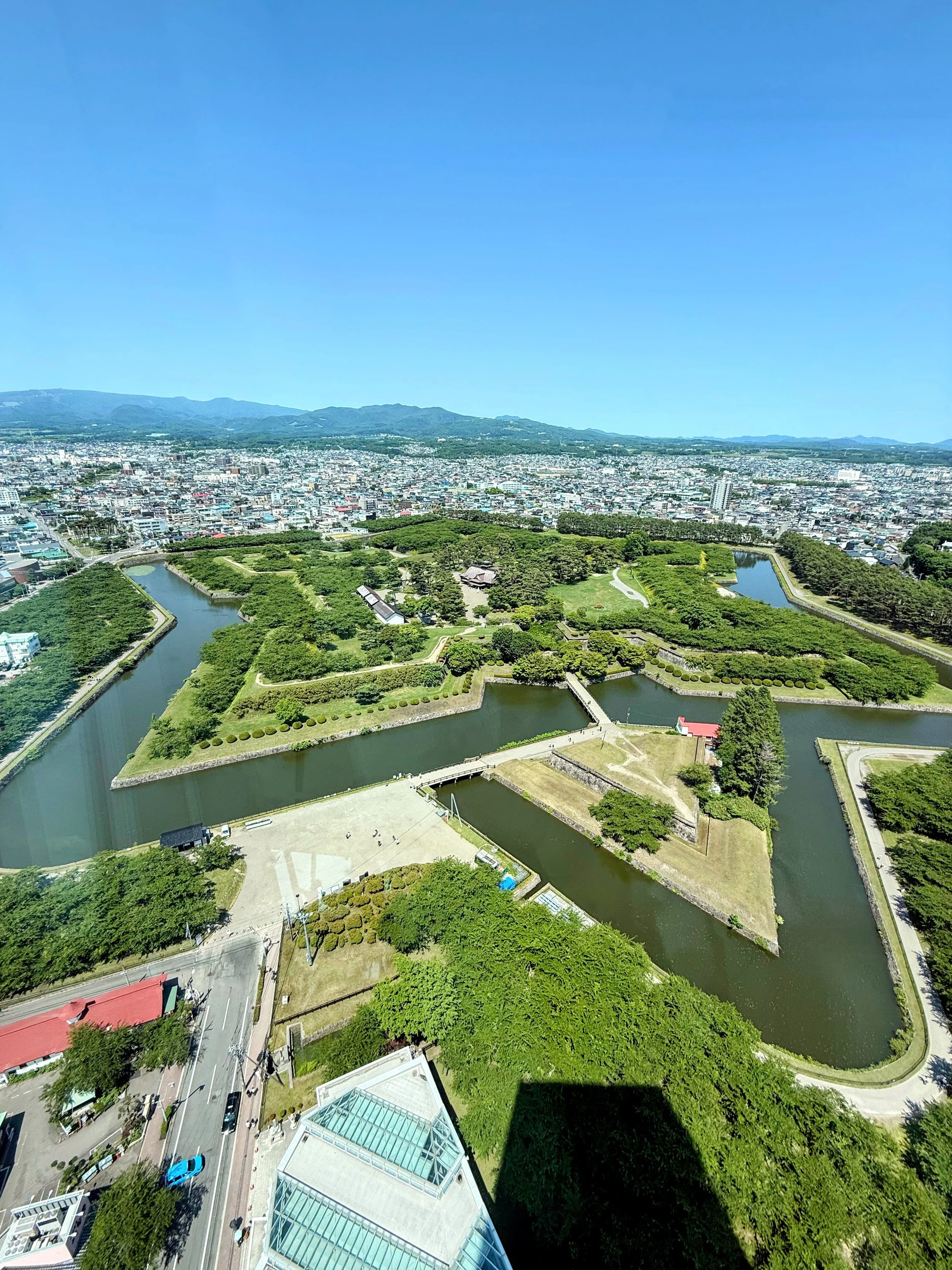 Aerial view of a historic Japanese castle surrounded by water moats, with a cityscape and mountains in the background under a clear blue sky.