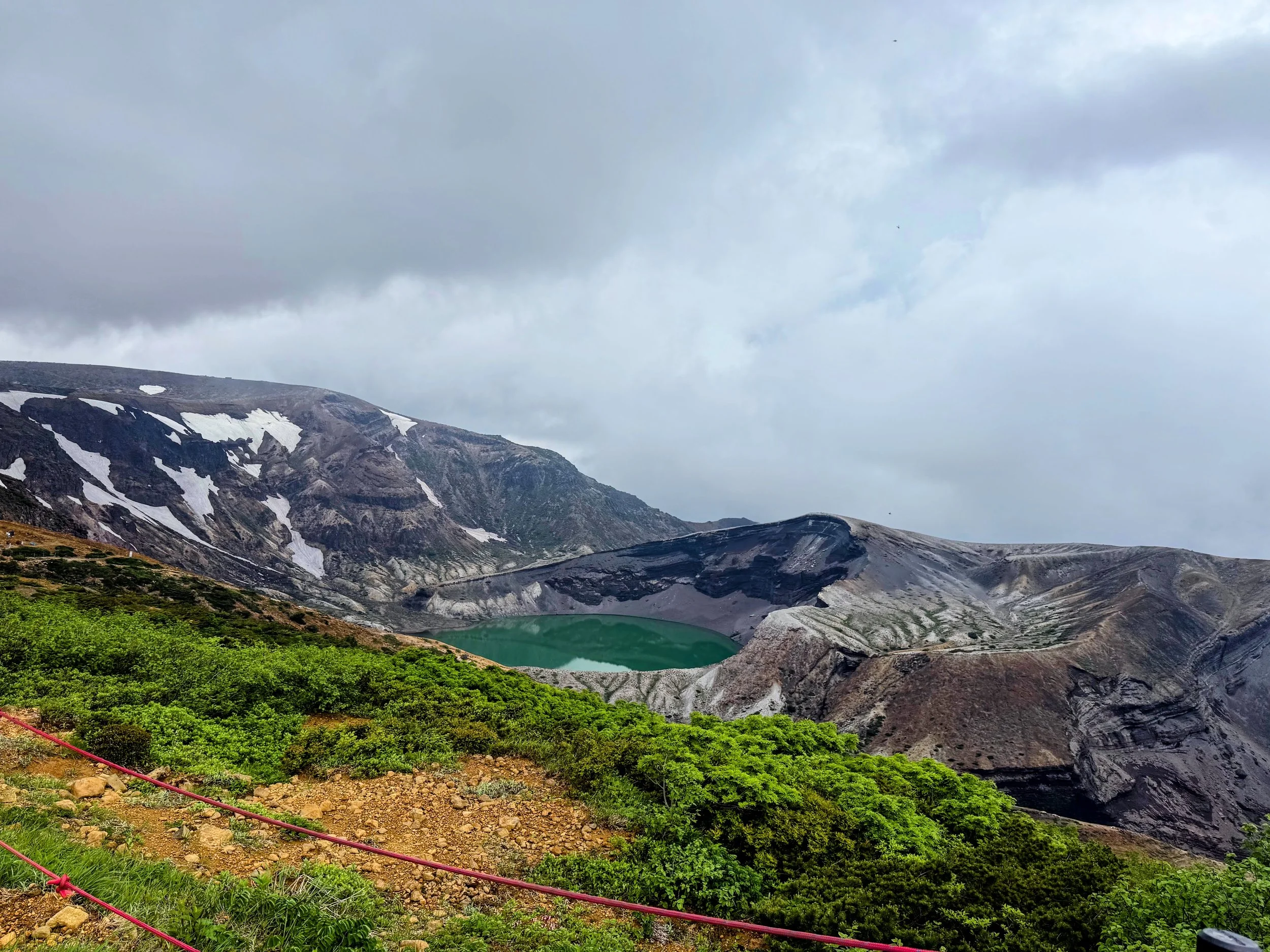 A mountainous landscape with snow patches on dark rocky slopes, a small greenish crater lake, and lush green vegetation in the foreground under a cloudy sky.